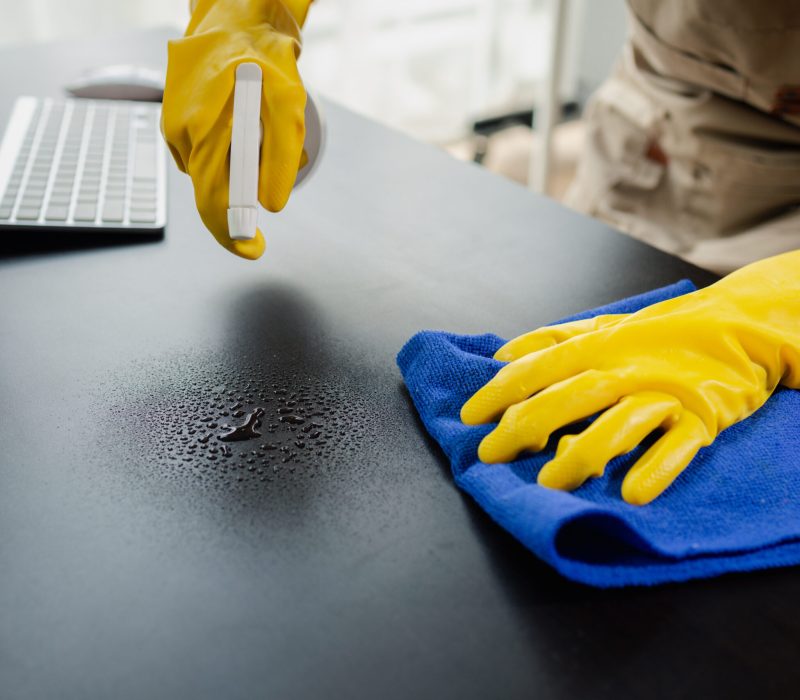 Cleaning desk surface in office with sanitizer spray, wear gloves and wipe the table with a towel, the housekeeper is cleaning the work desk for hygiene because of the Covid-19, cleaning idea.