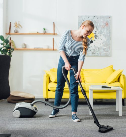 Smiling woman cleaning carpet with vacuum cleaner in living room